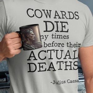 May include: A gray t-shirt with the text "Cowards Die many times before their ACTUAL DEATHS - Julius Caesar" printed on it. A person is holding a black mug with a picture of Julius Caesar on it.