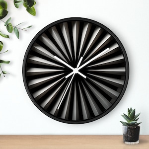 May include: A modern black and white wall clock with a radial design. The clock features a black frame and white clock hands. The face of the clock has a pattern of black and white angled lines. A small potted succulent is visible in the lower right corner.