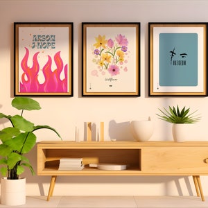 May include: Three framed prints on a white wall above a wooden shelf. The left print features a pink and orange flame with the text "arson j-hope". The middle print features a cluster of pink, yellow, and purple wildflowers with the text "Wildflower". The right print features a blue background with a black silhouette of a face and the text "Haegeum".