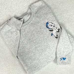 Könnte beinhalten: Ein hellgraues Sweatshirt mit Rundhalsausschnitt und langen Ärmeln. Auf der oberen linken Brust befindet sich eine Cartoon-Teufel-Grafik in Blau, Schwarz und Weiß. Das Ärmelbündchen hat ein blaues Herz und den Buchstaben "N".