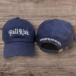 May include: Two navy blue baseball caps. One cap has the words "Fall Risk" embroidered in white script. The other cap has the phrase "If you can read this pick me up" embroidered in white. Both caps have a distressed look.