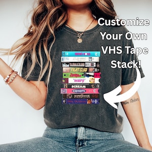 Puede incluir: Una camiseta gris oscuro con una pila de cintas VHS con títulos de películas como "Fight Club" y "Scream". La camiseta tiene el texto blanco "Customize Your Own VHS Tape Stack!". La persona lleva vaqueros azules y zapatos blancos.