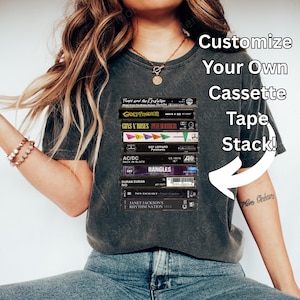 May include: Charcoal grey t-shirt displaying a collection of cassette tapes, including bands such as Guns N' Roses and AC/DC. The shirt has the text "Customize Your Own Cassette Tape Stack!" in white. The model is wearing blue jeans and white shoes.