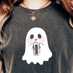May include: Dark gray t-shirt featuring a white ghost graphic with pink cheeks, holding a drink with a straw. The drink has the word "Custom" printed on it. The shirt is a comfortable, casual style.