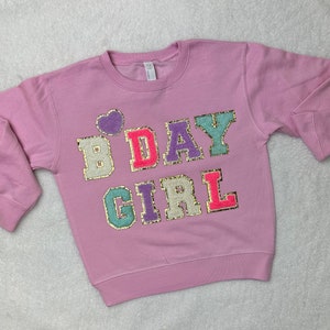 May include: A lavender sweatshirt with the words "B DAY GIRL" in colorful, glittery letters. The letters are outlined in gold.
