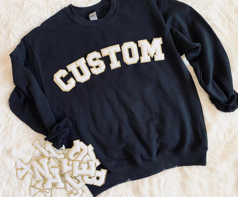 May include: A black long-sleeve crewneck sweatshirt featuring the word CUSTOM in large, white, block-style letters with gold glitter trim. A pile of additional individual white and gold glitter letter patches is positioned at the bottom left.