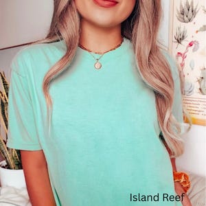 May include: A light green t-shirt with the text "Island Reef" at the bottom. The shirt is worn with a gold necklace. The model has long blonde hair.