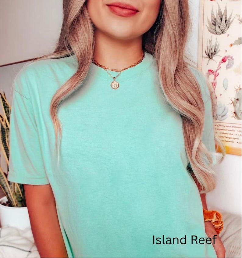 May include: A light green t-shirt with the text "Island Reef" at the bottom. The shirt is worn with a gold necklace. The model has long blonde hair.