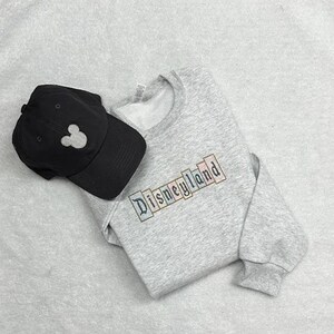 K&ouml;nnte beinhalten: Ein graues Sweatshirt mit dem Wort "Disneyland" in bunten Buchstaben gestickt, kombiniert mit einer schwarzen Baseballkappe mit einer wei&szlig;en Mickey Mouse Silhouette. Die Artikel liegen auf einer hellgrauen Oberfl&auml;che.