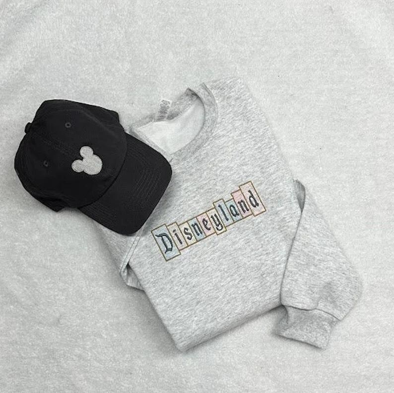 K&ouml;nnte beinhalten: Ein graues Sweatshirt mit dem Wort "Disneyland" in bunten Buchstaben gestickt, kombiniert mit einer schwarzen Baseballkappe mit einer wei&szlig;en Mickey Mouse Silhouette. Die Artikel liegen auf einer hellgrauen Oberfl&auml;che.