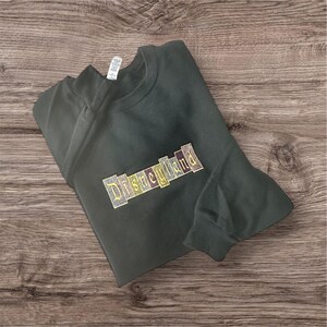 K&ouml;nnte beinhalten: Dunkelgr&uuml;nes Sweatshirt mit dem gestickten Wort "Disneyland" in einem farbenfrohen Blockbuchstaben-Design. Das Sweatshirt liegt gefaltet auf einer Holzoberfl&auml;che.