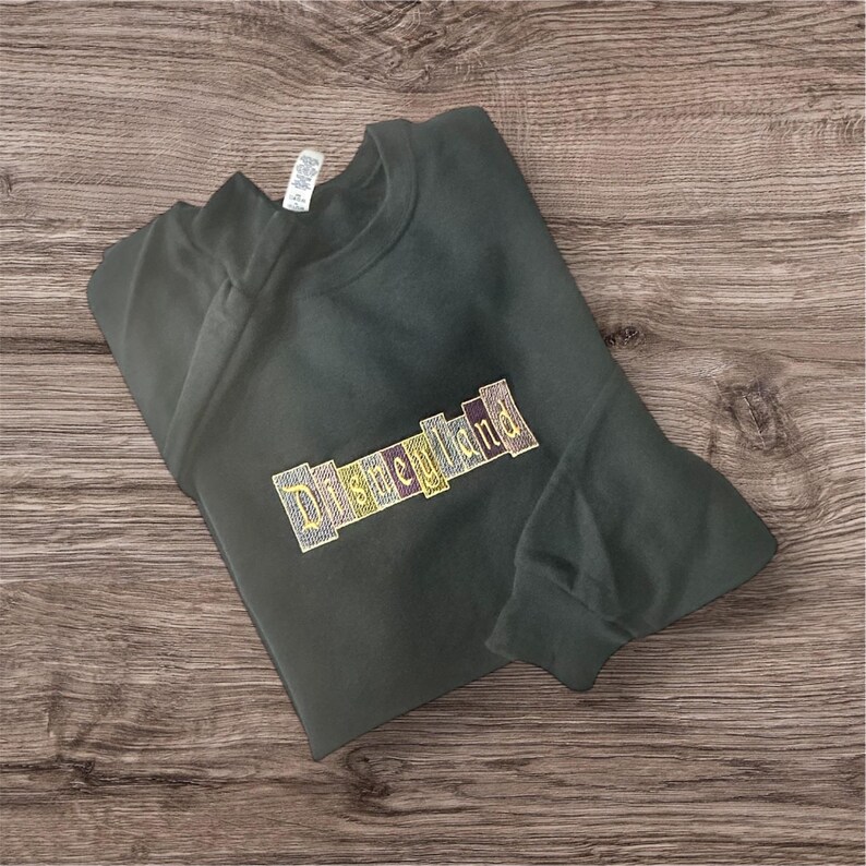 K&ouml;nnte beinhalten: Dunkelgr&uuml;nes Sweatshirt mit dem gestickten Wort "Disneyland" in einem farbenfrohen Blockbuchstaben-Design. Das Sweatshirt liegt gefaltet auf einer Holzoberfl&auml;che.