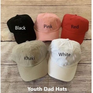 May include: Five youth dad hats in different colors: black, pink, red, khaki, and white. The hats are arranged in a semi-circle on a wooden surface. The text "Youth Dad Hats" is printed below the hats.