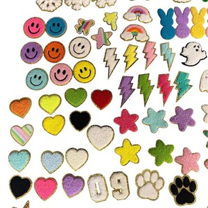 May include: A collection of assorted chenille patches featuring various shapes including smiley faces, hearts, stars, rainbows, lightning bolts, snowflakes, numbers 0 and 9, paw prints, and a ghost, all with gold-toned trim edges.