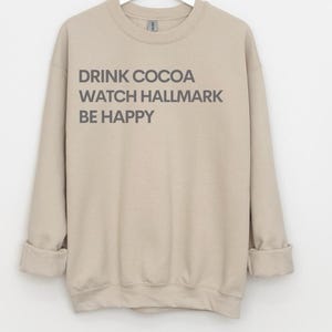 Könnte beinhalten: Beiges Sweatshirt mit dem grauen Text "DRINK COCOA WATCH HALLMARK BE HAPPY". Die langen Ärmel sind an den Handgelenken umgeschlagen. Das Sweatshirt besteht aus einem weichen, bequemen Material.