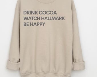 May include: A beige sweatshirt with the text "DRINK COCOA WATCH HALLMARK BE HAPPY" in gray lettering. The long sleeves are cuffed at the wrists. The sweatshirt is made of a soft, comfortable material.