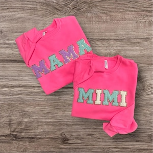 May include: Two bright neon pink crewneck sweatshirts. One sweatshirt features the word MAMA in large, glittery, multi-colored block letters. The other sweatshirt features the word MIMI in large, glittery, multi-colored block letters.
