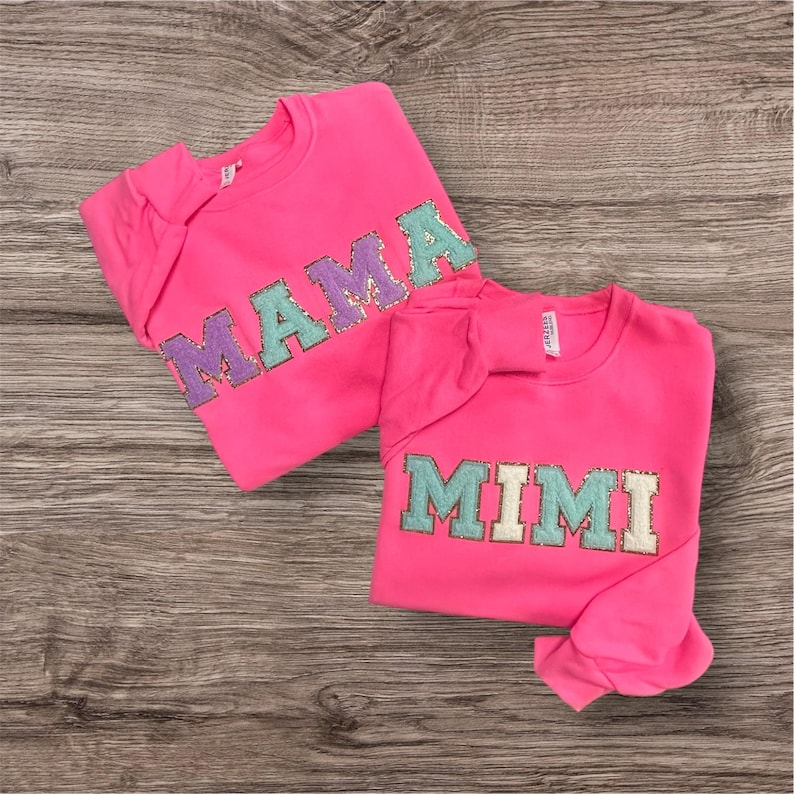 May include: Two bright neon pink crewneck sweatshirts. One sweatshirt features the word MAMA in large, glittery, multi-colored block letters. The other sweatshirt features the word MIMI in large, glittery, multi-colored block letters.