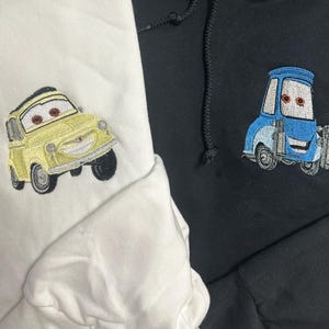 May include: Two hoodies, one white and one black, each embroidered with a cartoon car design. The white hoodie features a yellow car, while the black hoodie displays a blue car. Both designs have expressive eyes.