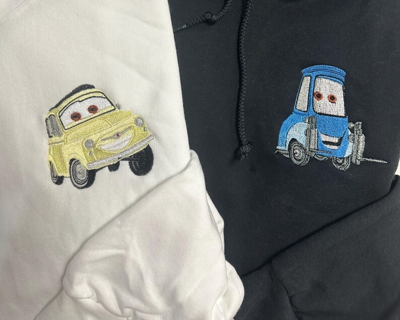 May include: Two hoodies, one white and one black, each embroidered with a cartoon car design. The white hoodie features a yellow car, while the black hoodie displays a blue car. Both designs have expressive eyes.