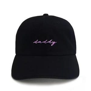 Puede incluir: Gorra de béisbol negra con visera curva. La palabra "daddy" está bordada en cursiva, en color morado claro en la parte delantera. La gorra está hecha de una tela suave y tiene una corona redondeada.