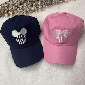 Könnte beinhalten: Zwei Baseballkappen, eine marineblaue und eine rosa, werden gezeigt. Jede Kappe hat ein weißes Mickey Mouse-Kopf-Design mit einem Monogramm. Die rosa Kappe hat eine Schleife auf den Ohren. Die Kappen befinden sich auf einer weißen Oberfläche.
