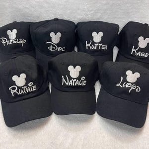 Puede incluir: Una colección de gorras de béisbol negras con nombres bordados en blanco y un diseño de cabeza de Mickey Mouse. Los nombres incluyen Presley, Doc, Kutter, Kase, Ruthie, Natalie y Lloyd.