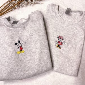 Puede incluir: Dos sudaderas de color gris claro con personajes de dibujos animados bordados. Una presenta a Mickey Mouse en negro, rojo, amarillo y blanco. La otra tiene a Minnie Mouse con un vestido de lunares rojos y blancos. Ambas sudaderas están dobladas.
