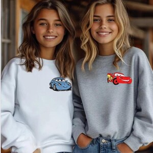 May include: Two sweatshirts, one white and one gray, each featuring a cartoon car design. The white sweatshirt has a blue car, while the gray one displays a red car. Both sweatshirts are paired with blue jeans.