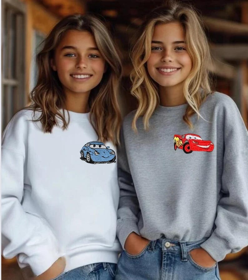 May include: Two sweatshirts, one white and one gray, each featuring a cartoon car design. The white sweatshirt has a blue car, while the gray one displays a red car. Both sweatshirts are paired with blue jeans.