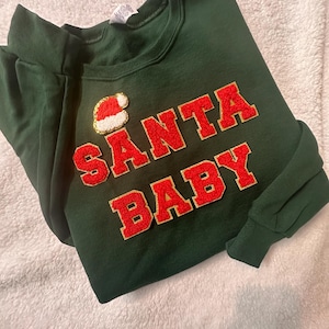 May include: A dark green long-sleeve sweatshirt featuring the text SANTA BABY in red varsity-style letters with gold glitter trim. Above the text is a small patch of a red and white Santa hat. The sweatshirt has a crew neckline.