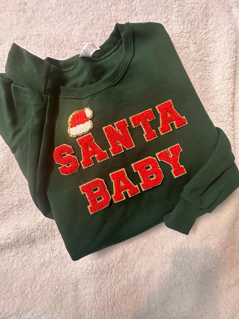 May include: A dark green long-sleeve sweatshirt featuring the text SANTA BABY in red varsity-style letters with gold glitter trim. Above the text is a small patch of a red and white Santa hat. The sweatshirt has a crew neckline.