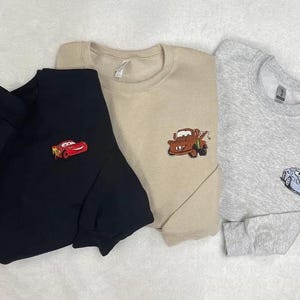 May include: Three crewneck sweatshirts in black, tan, and gray, each embroidered with a different character from the animated movie Cars. The black sweatshirt features Lightning McQueen, the tan one has Mater, and the gray one has a blue car.