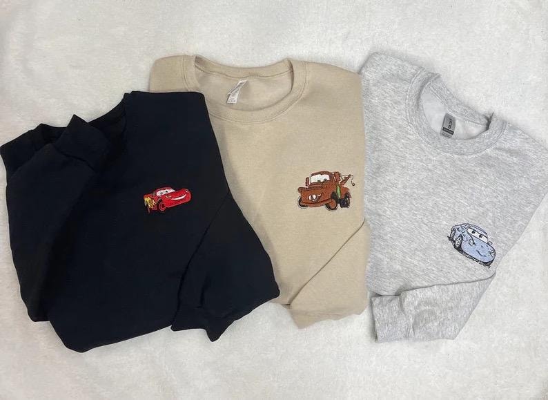 May include: Three crewneck sweatshirts in black, tan, and gray, each embroidered with a different character from the animated movie Cars. The black sweatshirt features Lightning McQueen, the tan one has Mater, and the gray one has a blue car.