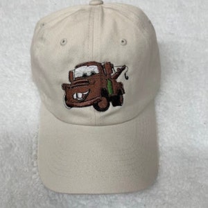 May include: A beige baseball cap with an embroidered image of Mater from the Disney Pixar movie Cars. The image shows Mater's face and his tow truck.