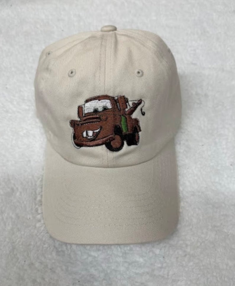 May include: A beige baseball cap with an embroidered image of Mater from the Disney Pixar movie Cars. The image shows Mater's face and his tow truck.