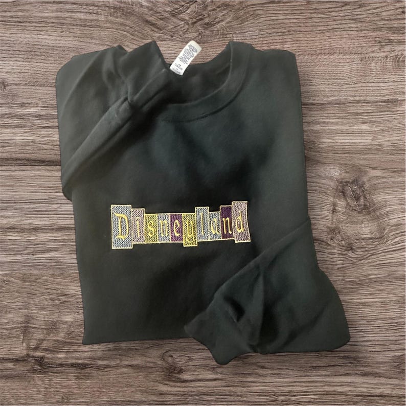 K&ouml;nnte beinhalten: Dunkelgr&uuml;nes Sweatshirt mit dem gestickten Text "Disneyland" in einem farbenfrohen Blockbuchstaben-Design. Das Sweatshirt ist gefaltet, wobei die &Auml;rmel und der Halsausschnitt sichtbar sind. Der Hintergrund ist eine Holzoberfl&auml;che.
