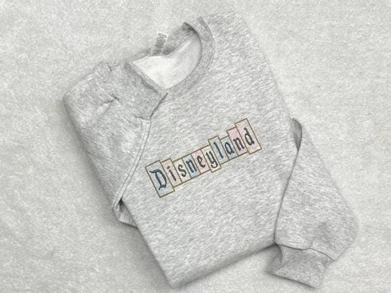 K&ouml;nnte beinhalten: Ein hellgraues Sweatshirt mit dem Wort "Disneyland" in einem farbenfrohen Blockbuchstaben-Design auf der Brust gestickt. Das Sweatshirt hat einen Rundhalsausschnitt und lange &Auml;rmel mit gerippten B&uuml;ndchen.