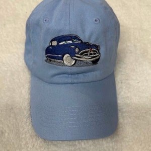 May include: A light blue baseball cap with an embroidered image of a blue car. The car has a white windshield and white tires.