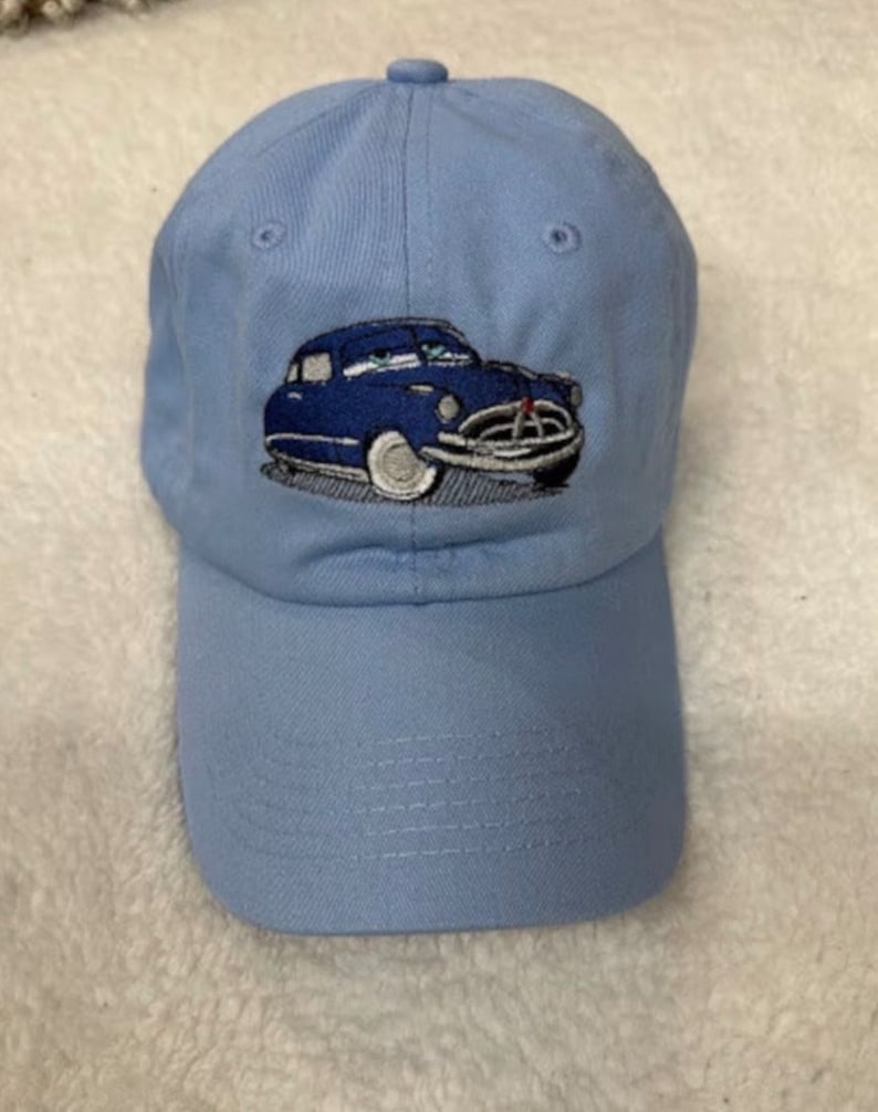 May include: A light blue baseball cap with an embroidered image of a blue car. The car has a white windshield and white tires.