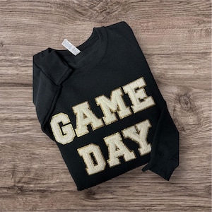 May include: A black long-sleeve sweatshirt featuring the text GAME DAY in large, white, textured varsity-style letters with a gold glitter outline. The garment has a crew neckline and a white brand tag visible at the inner collar area.