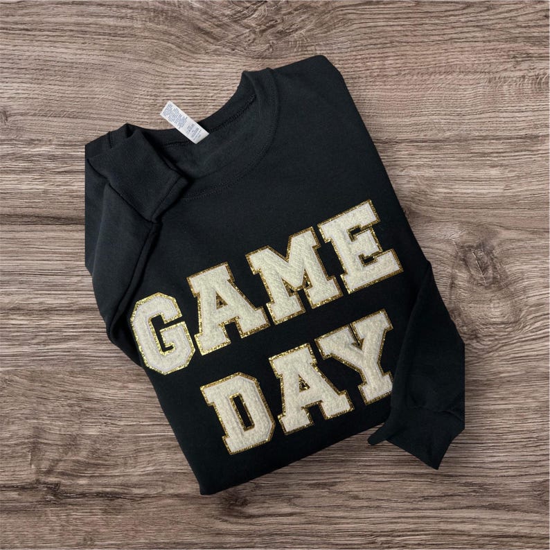 May include: A black long-sleeve sweatshirt featuring the text GAME DAY in large, white, textured varsity-style letters with a gold glitter outline. The garment has a crew neckline and a white brand tag visible at the inner collar area.