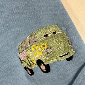 May include: A light blue sweatshirt featuring an embroidered green van with black wheels and floral accents. The van has a smiling face and is positioned in the lower right corner of the garment.