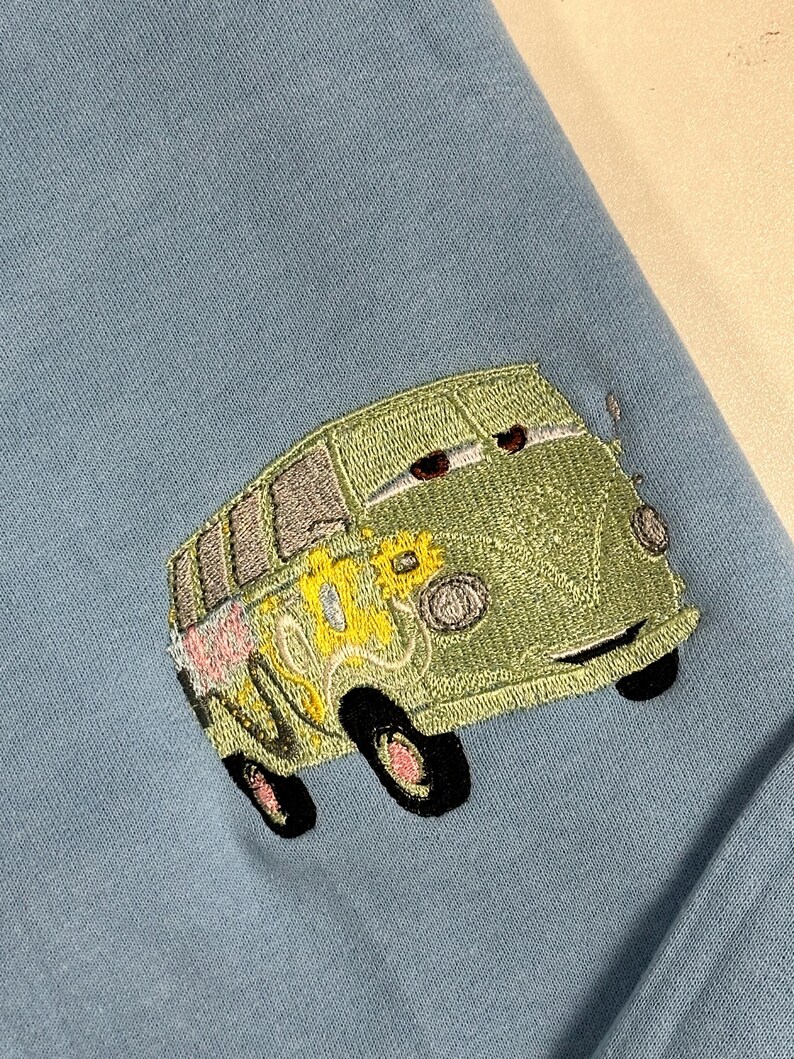 May include: A light blue sweatshirt featuring an embroidered green van with black wheels and floral accents. The van has a smiling face and is positioned in the lower right corner of the garment.