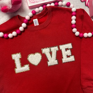 May include: A red long-sleeve sweatshirt featuring the word LOVE in white, textured, varsity-style letters with gold glitter outlines. The letter O is replaced by a white heart. A string of pink and white beads rests across the neckline.
