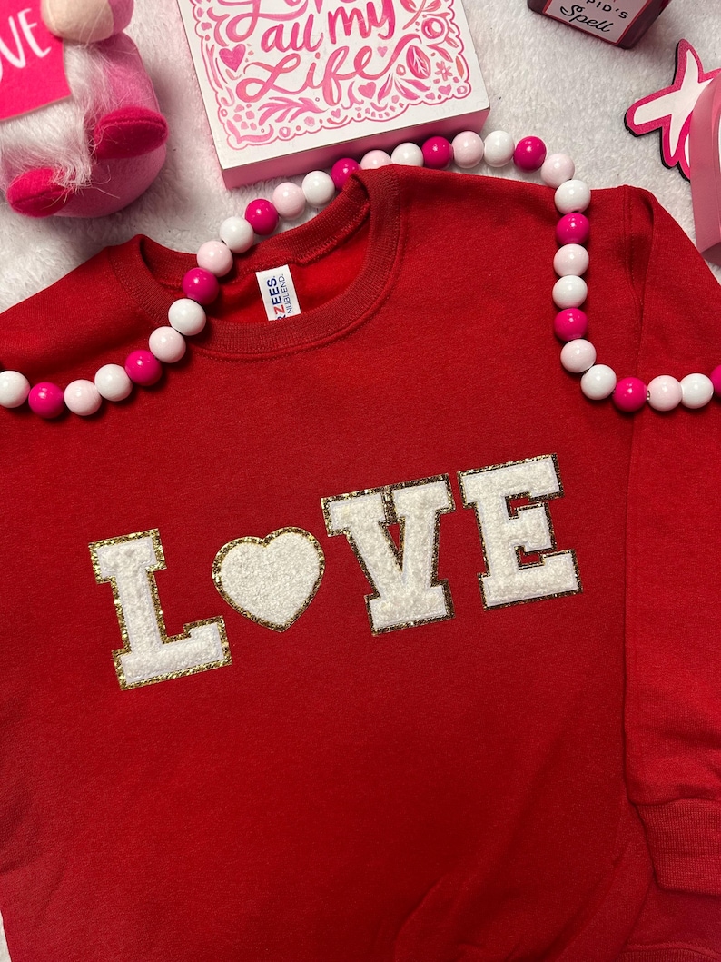 May include: A red long-sleeve sweatshirt featuring the word LOVE in white, textured, varsity-style letters with gold glitter outlines. The letter O is replaced by a white heart. A string of pink and white beads rests across the neckline.