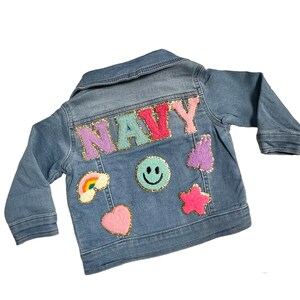 May include: Light blue denim jacket with the name "NAVY" in colorful, sequined letters across the back. The jacket features patches of a rainbow, smiley face, heart, star, and other shapes.