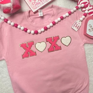 May include: A light pink crewneck sweatshirt featuring the letters X and O in bright pink chenille with gold glitter borders, separated by two white chenille hearts. The design is centered on the chest of the garment, which has a standard ribbed neckline.
