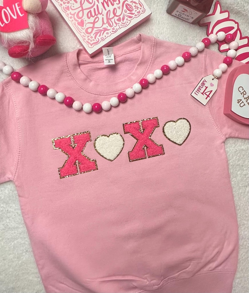 May include: A light pink crewneck sweatshirt featuring the letters X and O in bright pink chenille with gold glitter borders, separated by two white chenille hearts. The design is centered on the chest of the garment, which has a standard ribbed neckline.