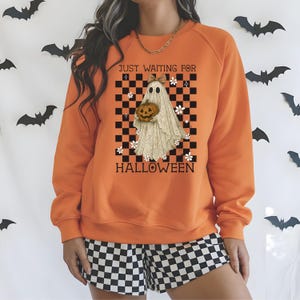May include: Orange sweatshirt with a Halloween-themed graphic. The graphic features a ghost holding a pumpkin against a black and white checkered background. The text "JUST WAITING FOR HALLOWEEN" is printed above and below the ghost.